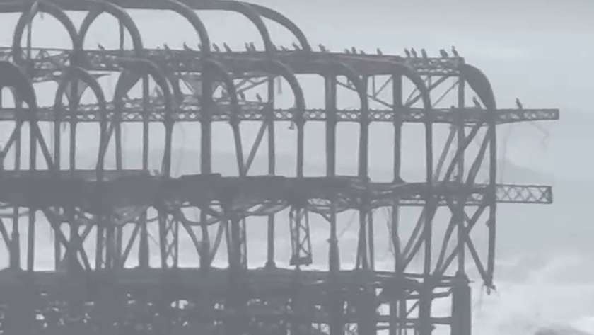 The arched iron frame of the West Pier with birds perched along the top and surf breaking around its base
