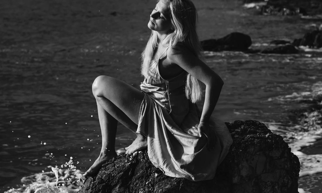 Black and white photograph of Tallulah Rendall sitting on a rock at the water’s edge, face tilted towards the light