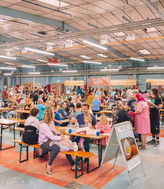 Visitors eating at long wooden tables in a large food hall with a high ceiling