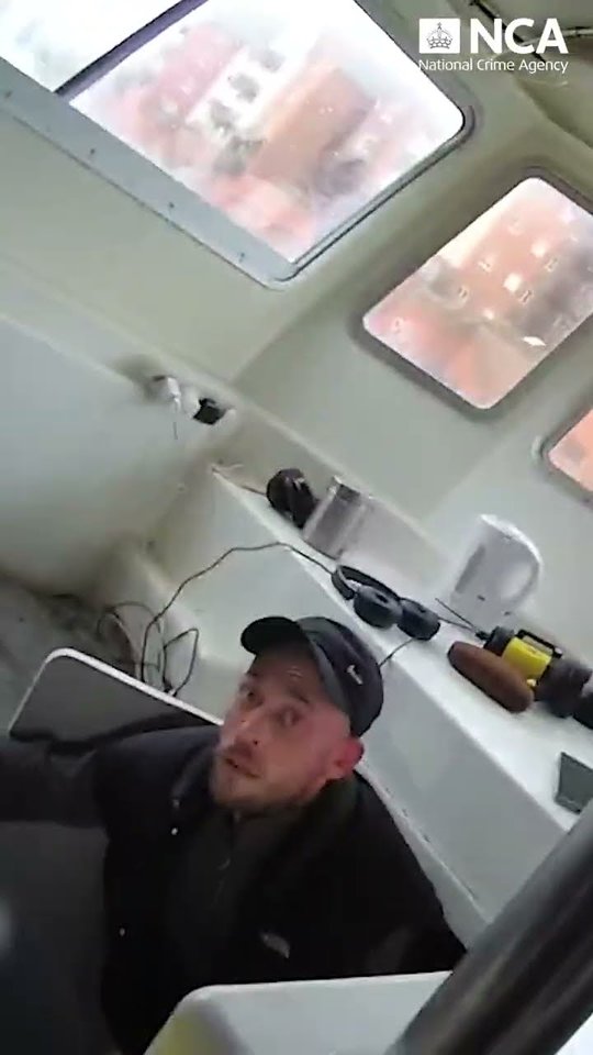 Still from National Crime Agency footage showing a man in a dark cap and jacket inside the cabin of a fishing vessel, with headphones and a kettle on a shelf behind him