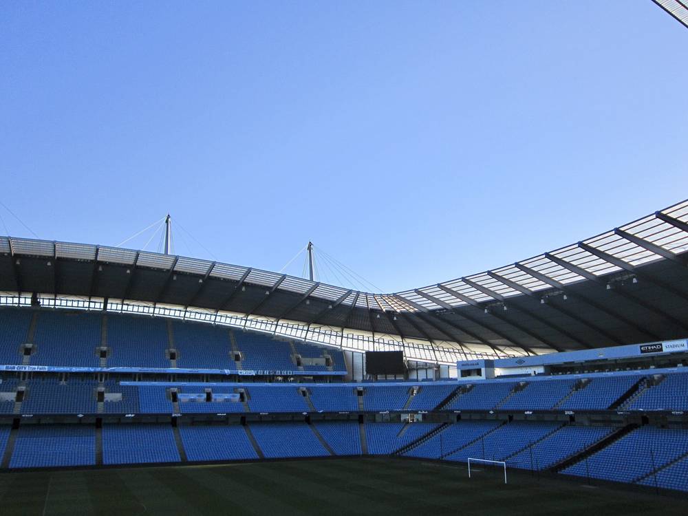 The blue seats and arched roof of a stand at the Etihad Stadium in Manchester under a clear sky