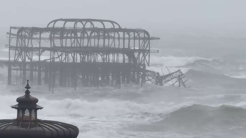 The West Pier frame in rough sea beyond the roof of the seafront bandstand, with large waves breaking in front of it