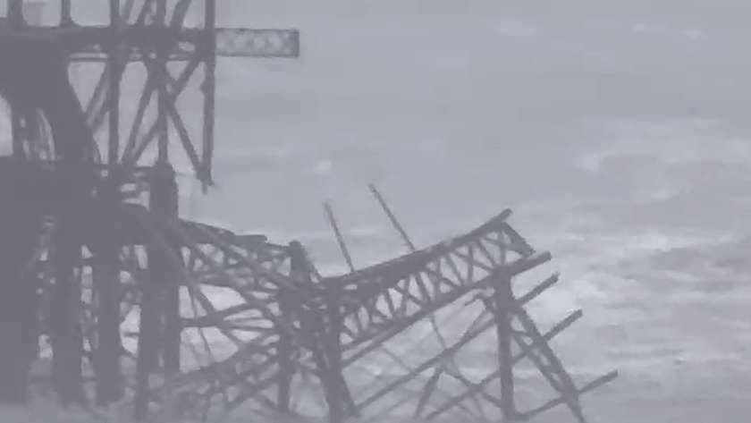 Close view of the West Pier ironwork with a tilted section of lattice girders lying in churning grey water at the base of the frame
