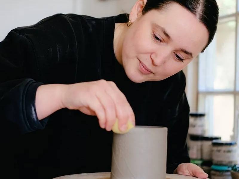A ceramicist smoothing the rim of a clay cylinder on a potter’s wheel in a bright studio