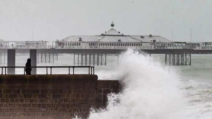 A large wave breaking over a sea wall on Brighton seafront with a pier in the grey distance and a person standing on the wall