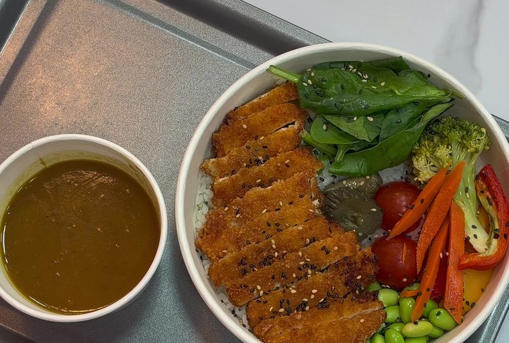 Chicken katsu curry in a white bowl with rice, spinach, carrot, tomato, broccoli and edamame, with a pot of curry sauce beside it