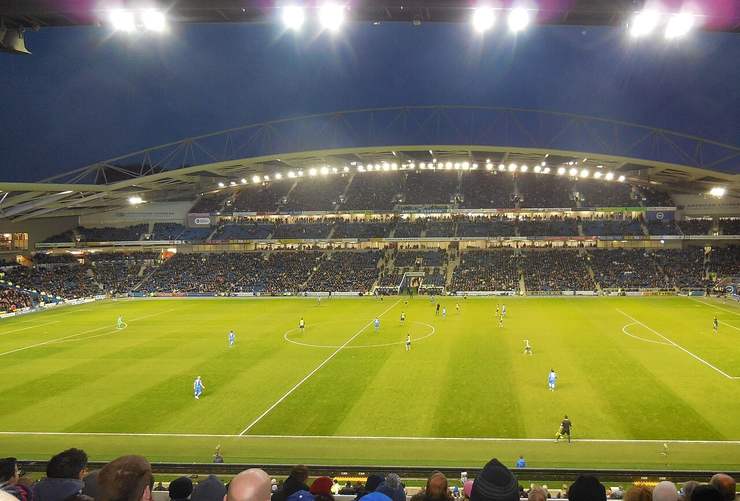 The Amex Stadium under floodlights during a match with the pitch lit green and the stands behind