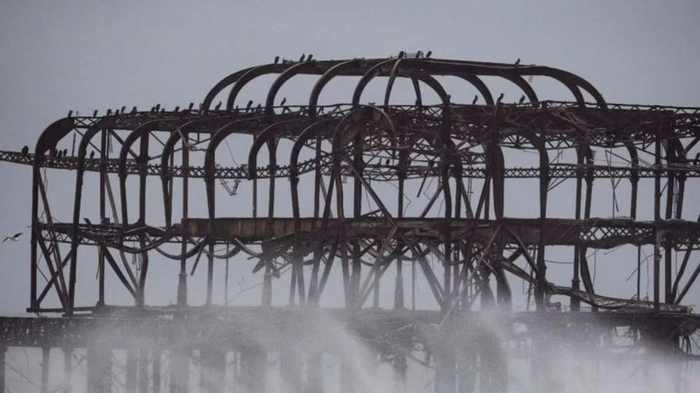 The curved iron arches of the West Pier standing above a large wave breaking in grey water