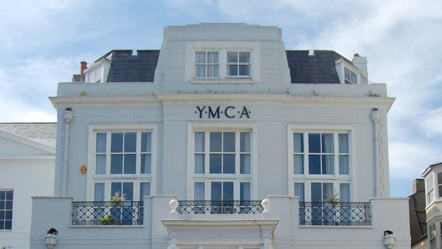 The upper floors of Steine House in Brighton, with the YMCA lettering, first-floor balconies and a slate mansard roof against a cloudy sky