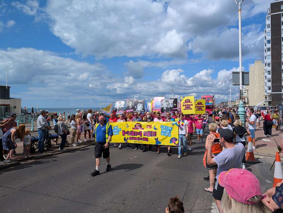 Marchers carry a bright yellow banner at the front of the Pride Community Parade on the Brighton seafront with crowds lining the road