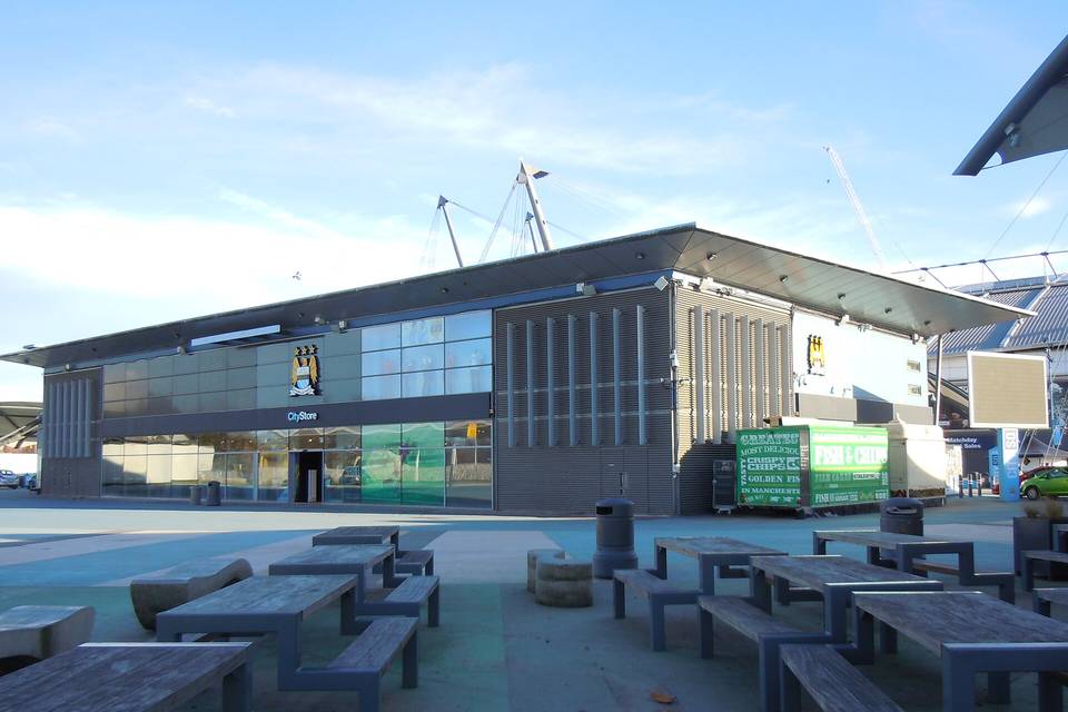 A glass-fronted building outside the Etihad Stadium in Manchester, with picnic benches in the foreground and a blue sky above