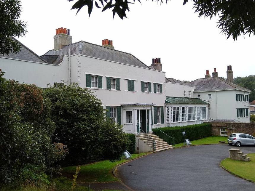 The white painted front of Preston Manor in Brighton with green shutters, tall chimneys and a curved brick wall beside a driveway and trees