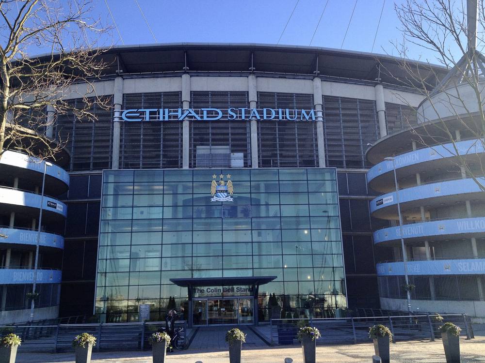 The glass-fronted Colin Bell Stand entrance of the Etihad Stadium in Manchester on a clear day, with the Etihad Stadium lettering above