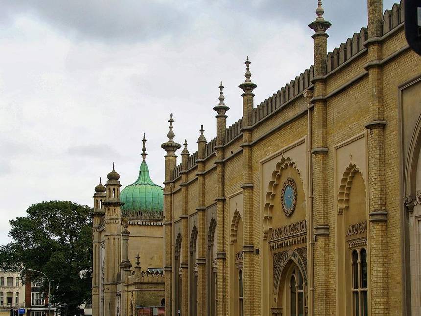 The long yellow brick wall of Brighton Museum and Art Gallery with Indian style arches and finials beside the green domed entrance gate of the Royal Pavilion on a cloudy day