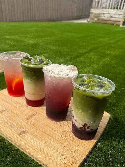 Four iced matcha and fruit drinks in clear cups on a wooden board on a lawn
