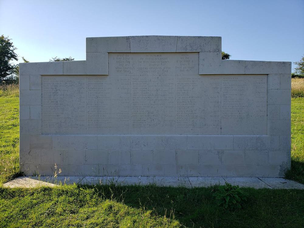 A pale stone memorial wall at the Chattri site with panels of names in English and Indian scripts, headed In honour of these soldiers of the Indian Army, in front of green grass and blue sky