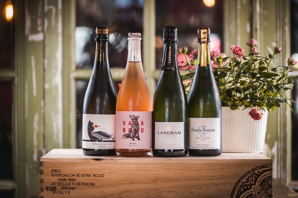 Four bottles of sparkling and still wine on a wooden crate in a window, with a pot of pink flowers beside them