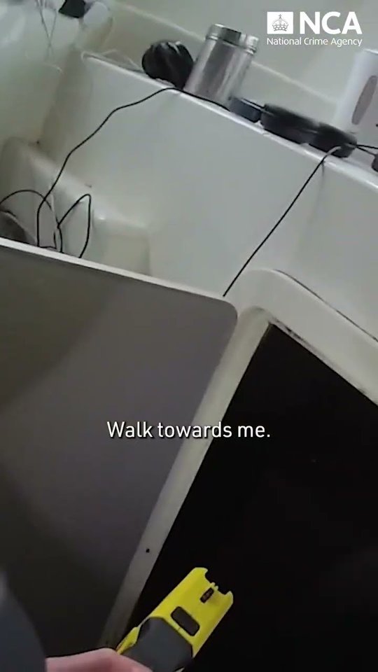 Still from National Crime Agency footage showing an officer holding a yellow Taser inside a vessel cabin, with the subtitle Walk towards me