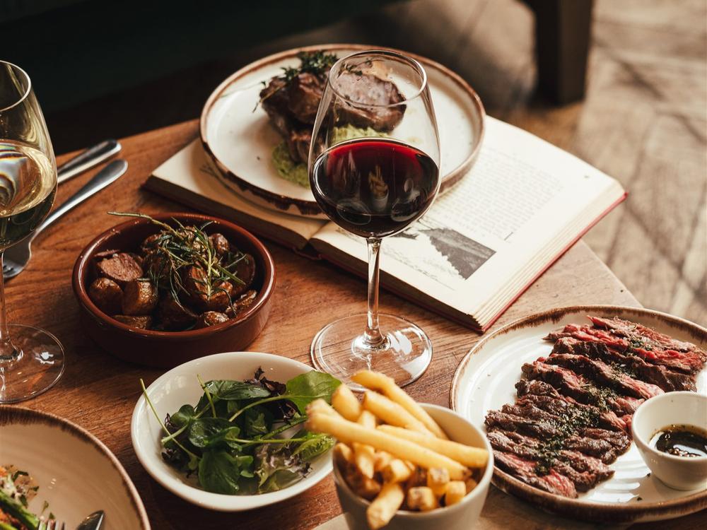 A table of small plates including salad, chips, sliced steak and a bowl of stew, with a glass of red wine and an open book