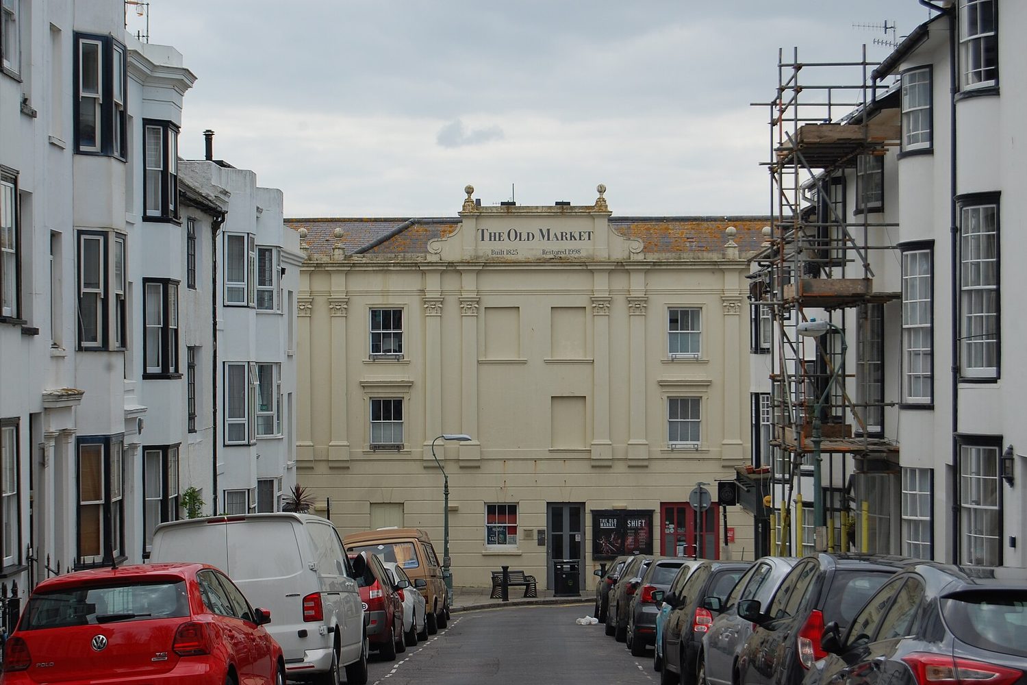 The cream coloured front of The Old Market in Hove, with the words The Old Market Built 1825 Restored 1998 on its pediment, at the end of a street of white terraced houses