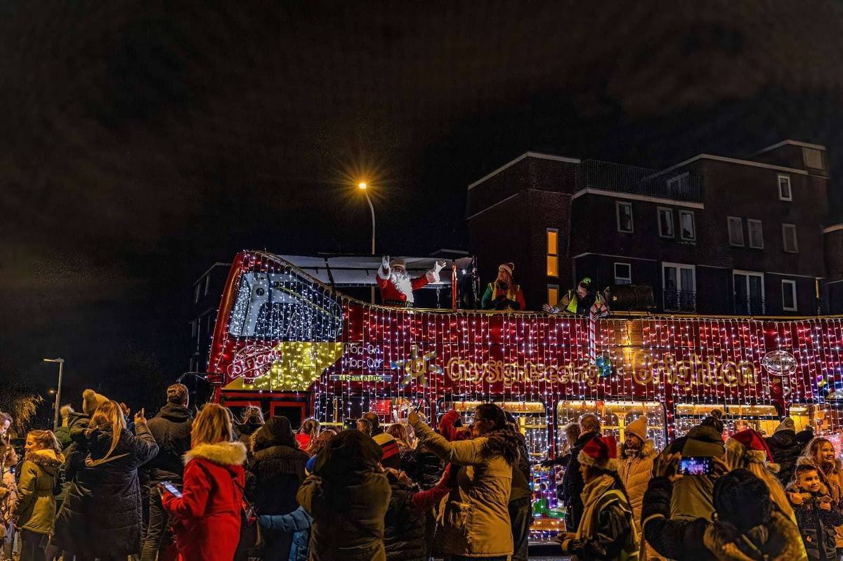 Children and parents in winter coats gather on a street at night to wave at the Santa Bus, a double decker covered in lights, with Santa and elves on the top deck