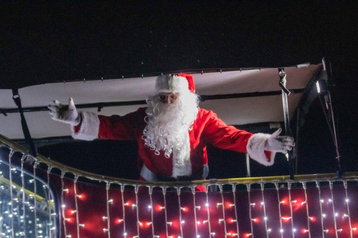 Santa Claus in a red suit and white beard standing on the top deck of the Santa Bus with both arms spread wide, in front of a curtain of red and white fairy lights at night