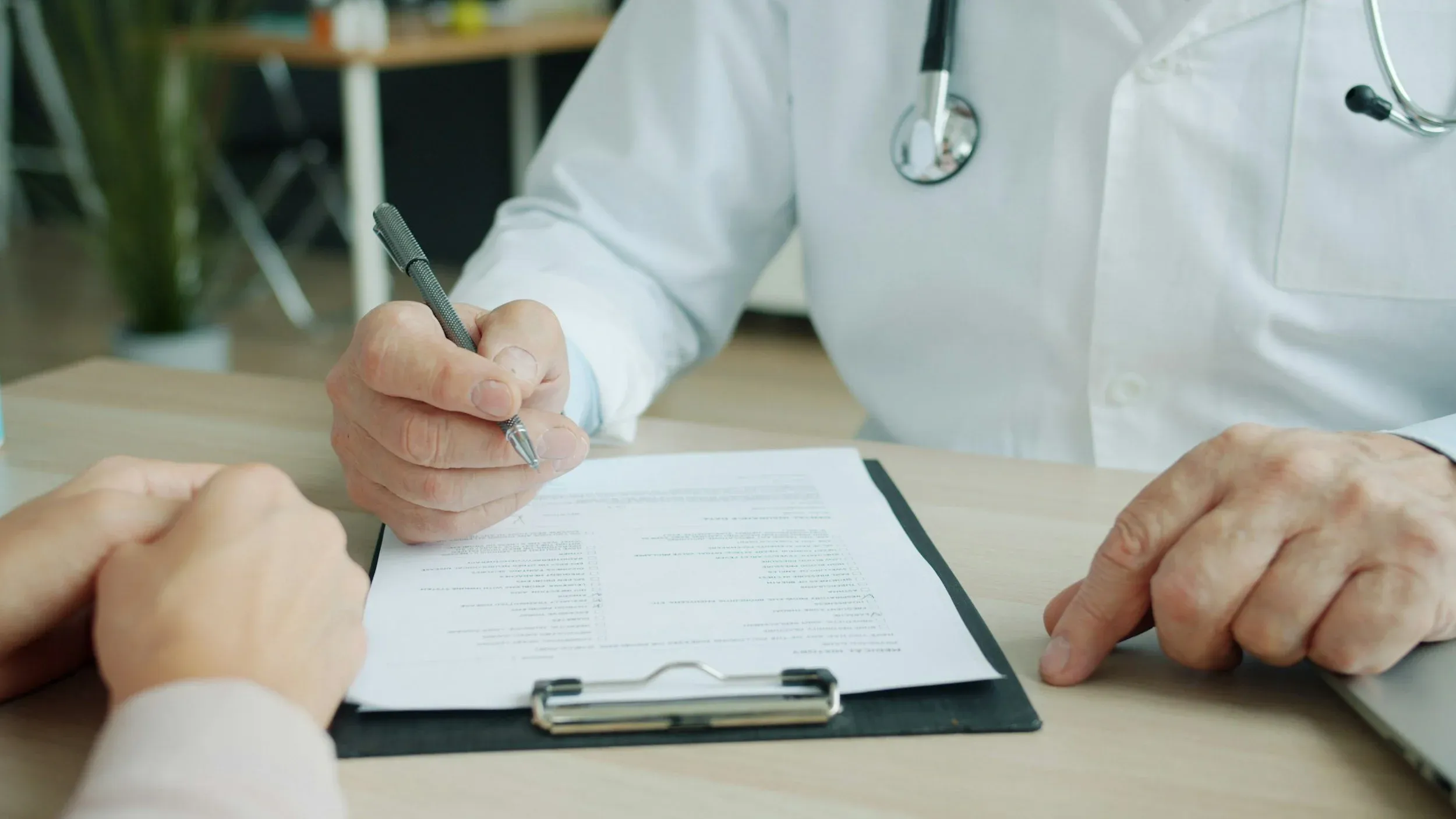 A researcher in a white coat completing paperwork on a clipboard with a participant seated across the desk