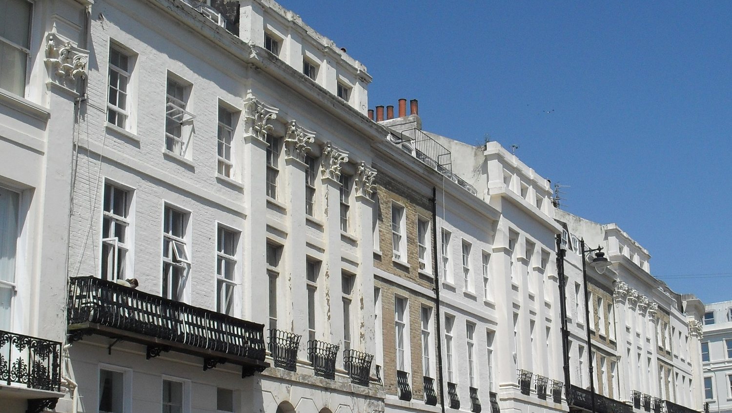 A terrace of white and cream stucco houses with black iron balconies running along a sunny street in Kemptown, Brighton, under a blue sky
