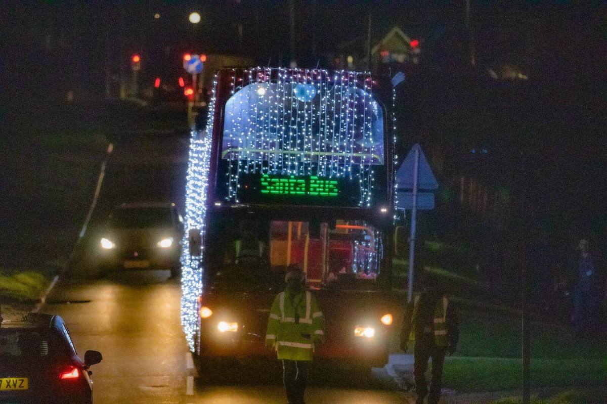 The Santa Bus approaching along a road at night, its front and top deck covered in white fairy lights and its destination screen reading Santa Bus, with stewards in yellow jackets standing nearby